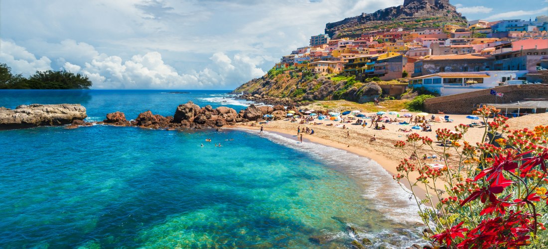 Blick auf den Strand von Castelsardo auf Sardinien mit klarem, blauem Meer und sanften Wellen. Im Hintergrund liegt die farbenfrohe Altstadt am Hang unterhalb einer Burg. Im Vordergrund blühen rote und orangefarbene Blumen, die der Szenerie zusätzliche Le