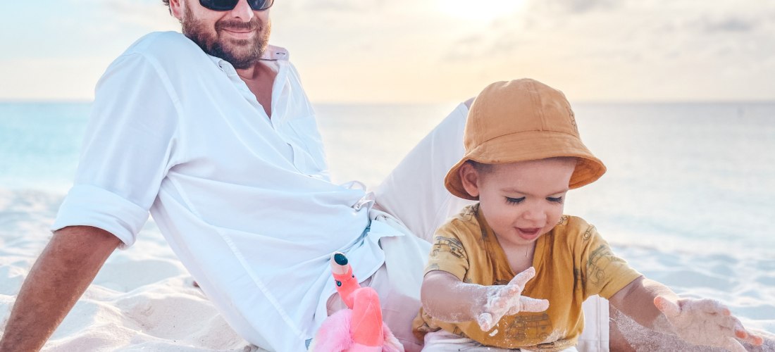 Ein Mann mit Sonnenbrille und weißem Hemd sitzt entspannt im weißen Sand am Strand von Aruba. Neben ihm spielt ein Kleinkind mit Sand, trägt einen beigefarbenen Sonnenhut und ein gelbes Shirt mit Auto-Motiven. Zwischen ihnen liegt ein pinkes Stoff-Flaming