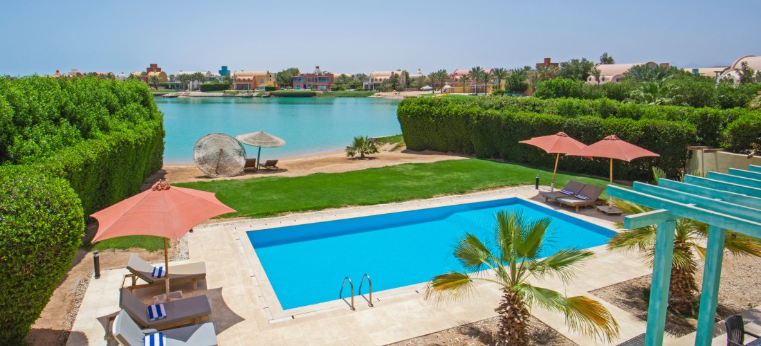 Private Poolanlage mit Sonnenliegen und Sonnenschirmen vor einer grünen Hecke. Dahinter liegt ein ruhiger türkisfarbener Wasserkanal mit Sandstrand und Schilf-Sonnenschirmen. Im Hintergrund bunte Ferienhäuser und Palmen unter klarem Himmel.