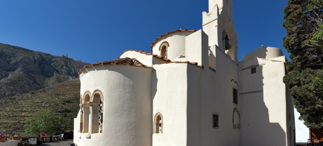 Außenansicht der byzantinischen Kirche Panagia Episkopi auf Santorini, Griechenland. Die Kirche ist weiß verputzt mit einem roten Ziegeldach und mehreren kleinen Kuppeln. Eine dekorative Glockenfassade mit zwei Glocken ragt in den klaren blauen Himmel. Im
