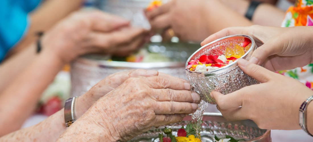 Das Bild zeigt eine thailändische Zeremonie, bei der junge Menschen mit silbernen, reich verzierten Schalen duftendes Wasser mit Blüten in die Hände älterer Menschen gießen. Diese Geste symbolisiert Respekt, Segnung und Dankbarkeit. Im Bild sind mehrere H