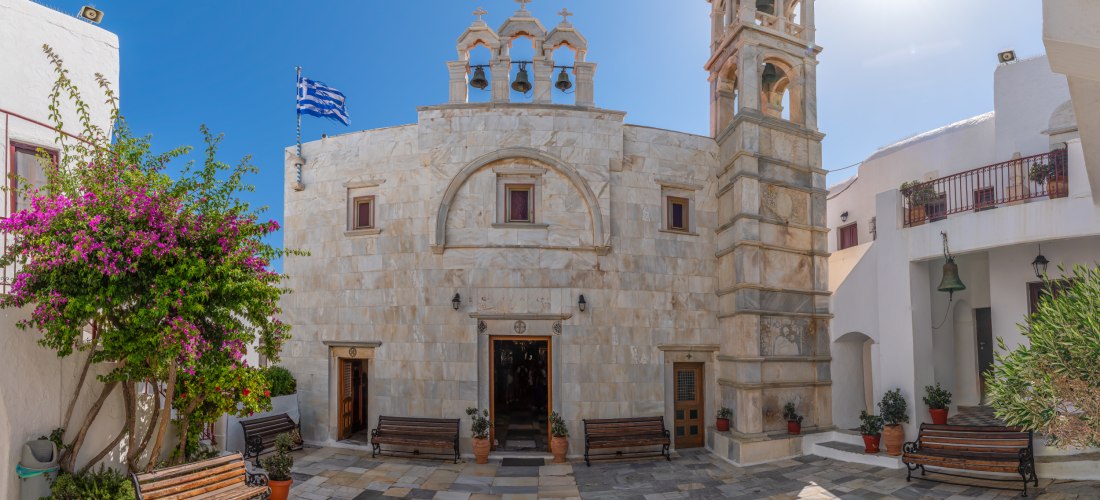 Heller Innenhof mit einer traditionellen griechischen Kirche aus hellem Stein. Über dem Eingang befinden sich drei Glocken unter kleinen Bögen. Rechts steht ein Glockenturm, im Hof blühen violette Bougainvillea-Blumen. Griechische Flagge weht im Hintergru