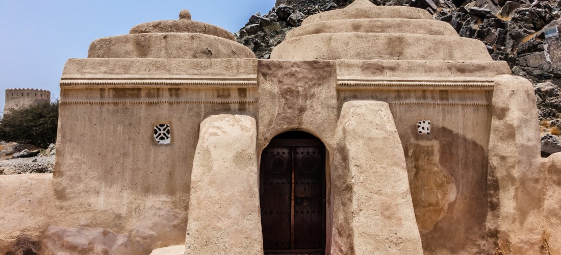 Frontansicht der Al-Bidyah-Moschee in Fujairah, Vereinigte Arabische Emirate. Historisches Lehmgebäude mit Kuppeln, traditioneller Eingangstür und Wachturm im Hintergrund.