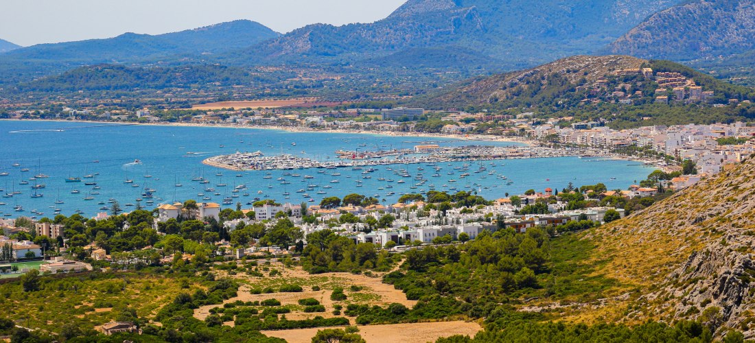 Panoramablick auf Port de Pollença im Norden Mallorcas. Zahlreiche Segelboote und Yachten liegen in der Bucht vor Anker, während im Hintergrund die imposante Serra de Tramuntana das Bild bestimmt. Die Stadt liegt eingebettet zwischen Hügeln und dem türkis
