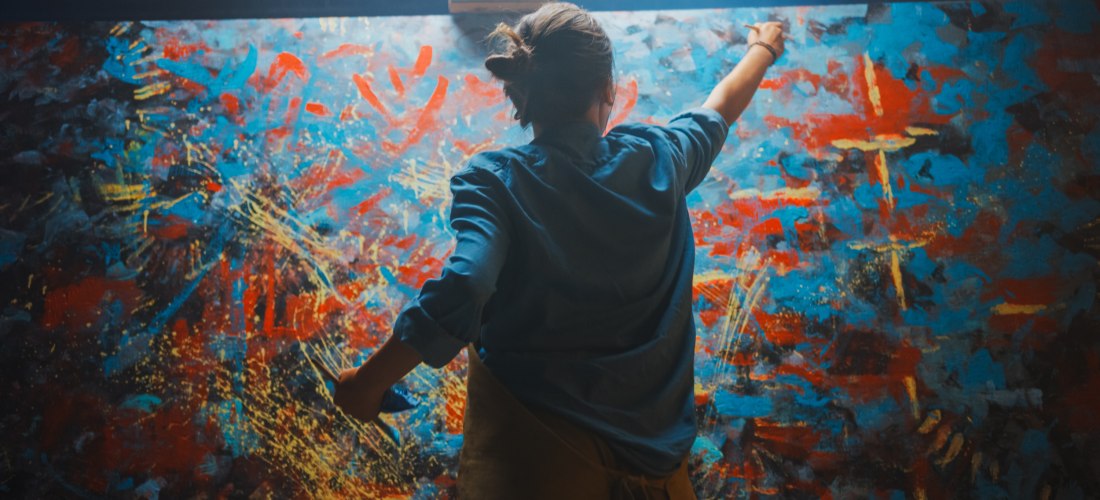 Eine Person von hinten in einem Atelier, die vor einer großen Leinwand steht und mit ausdrucksvollen Bewegungen ein farbenfrohes abstraktes Gemälde in Blau, Rot, Gelb und Schwarz malt.