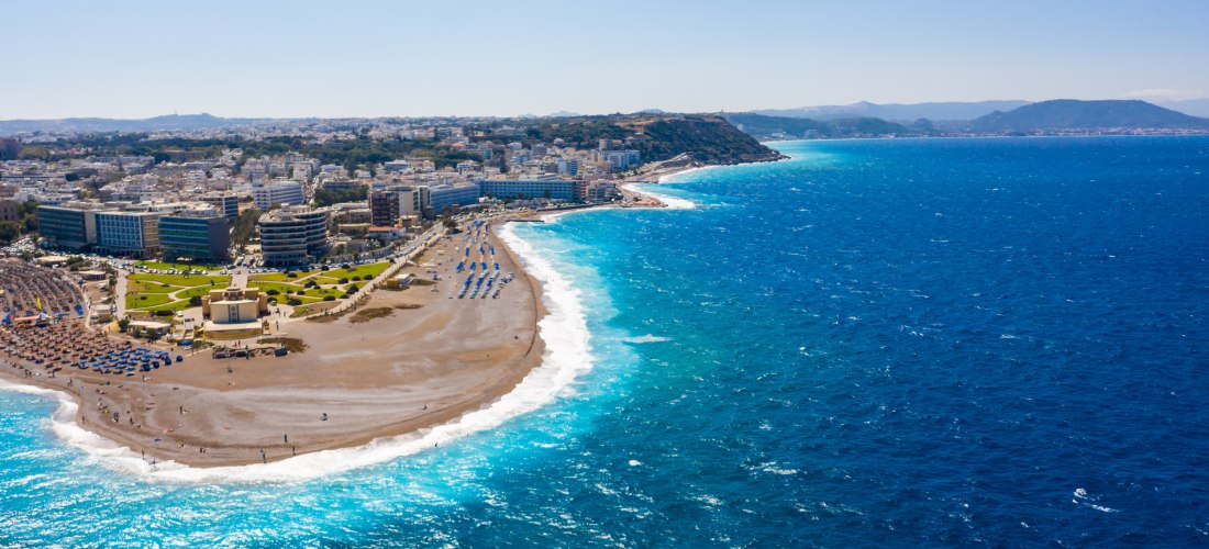 Die Aufnahme zeigt eine Panoramaansicht von Rhodos-Stadt auf der griechischen Insel Rhodos.   Der geschwungene Strand zieht sich entlang der Westküste, flankiert von einer breiten Promenade, gepflegten Grünanlagen und modernen Hotels.   Die Brandung des t