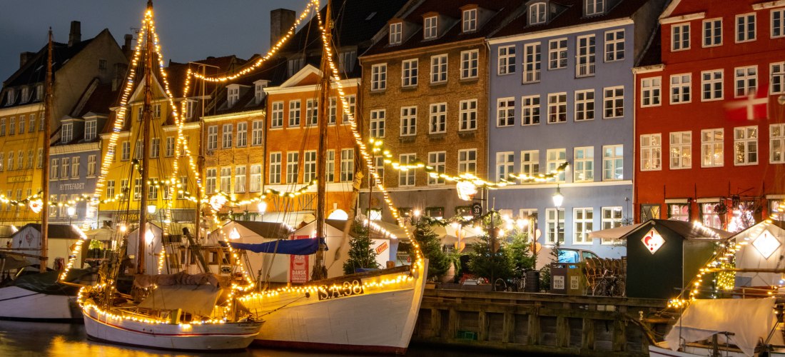 Abendliche Szene am Nyhavn in Kopenhagen. Bunte, dicht aneinander gereihte Häuser spiegeln sich im Wasser, davor liegen traditionelle Segelboote, die mit warm leuchtenden Lichterketten geschmückt sind. Eine gemütliche, winterliche Atmosphäre mit weihnacht