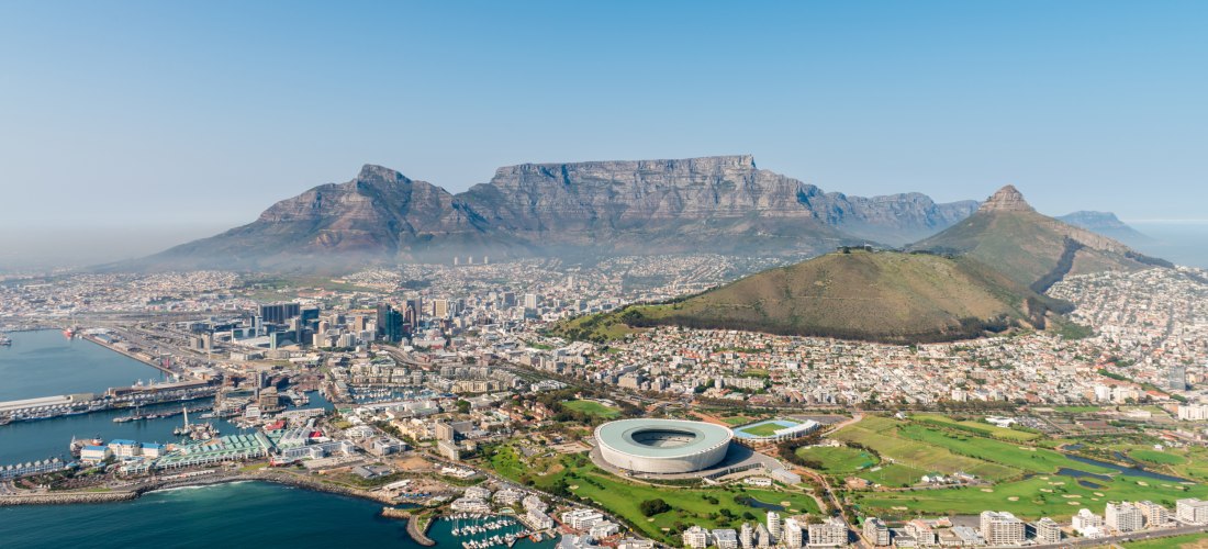 Luftaufnahme von Kapstadt in Südafrika. Im Vordergrund liegt der Atlantische Ozean mit Yachthafen und Uferpromenade. Zentral im Bild befindet sich das runde WM-Stadion von 2010. Dahinter erstreckt sich die Innenstadt mit zahlreichen Hochhäusern. Im Hinter