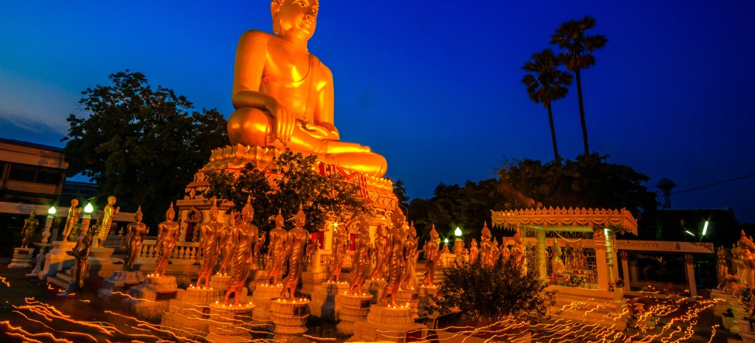 Das Bild zeigt eine eindrucksvoll beleuchtete, goldene Buddha-Statue bei Nacht. Sie ist von zahlreichen kleineren stehenden Buddhafiguren umgeben. Im Vordergrund verlaufen leuchtende Linien über den Boden – entstanden durch lange Belichtungszeit, während 
