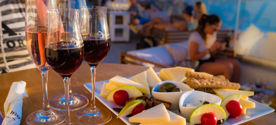 Drei Gläser Wein – zwei mit Rotwein und eines mit Rosé – stehen auf einem Tisch neben einer reich bestückten Käseplatte mit Apfel- und Birnenscheiben, Tomaten, Oliven und Crackern. Im Hintergrund sitzen Menschen auf Lounge-Möbeln auf einer Terrasse mit Bl