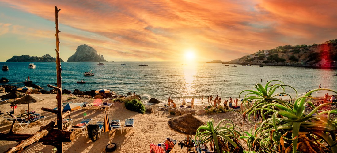 Ein malerischer Sonnenuntergang an einem Strand auf Ibiza. Der Himmel ist in leuchtenden Orange- und Rottönen gefärbt. Im Vordergrund stehen Sonnenliegen und Strohrundschirme auf dem Sand. Menschen stehen am Ufer oder schwimmen im Meer. Im Hintergrund rag