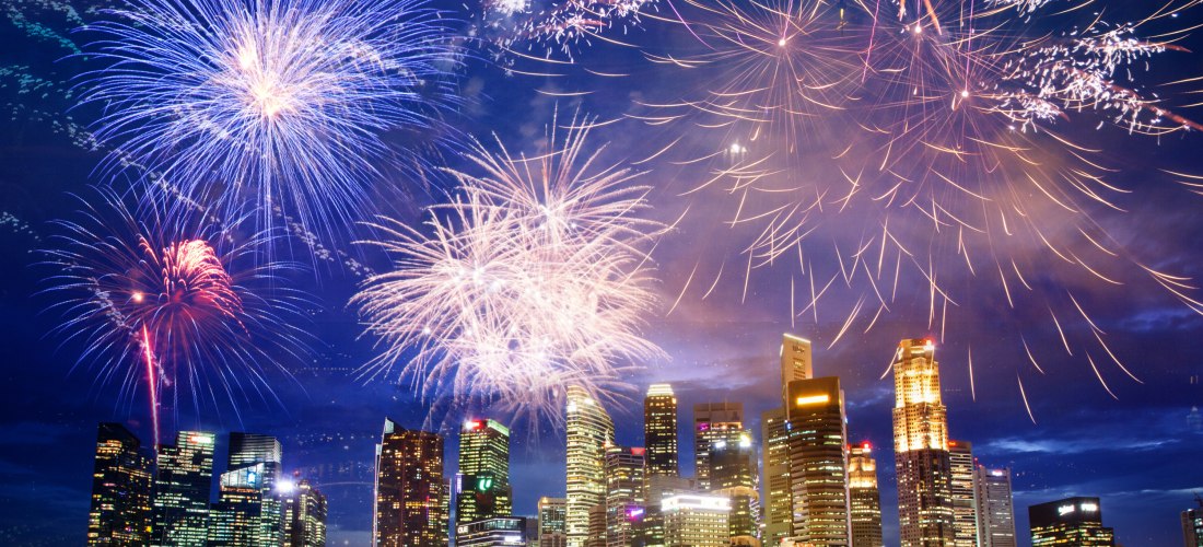 Feuerwerk über der nächtlichen Skyline von Singapur mit ihren glitzernden Hochhäusern. Die farbenfrohen Lichter des Feuerwerks spiegeln sich im Wasser und schaffen eine festliche Atmosphäre
