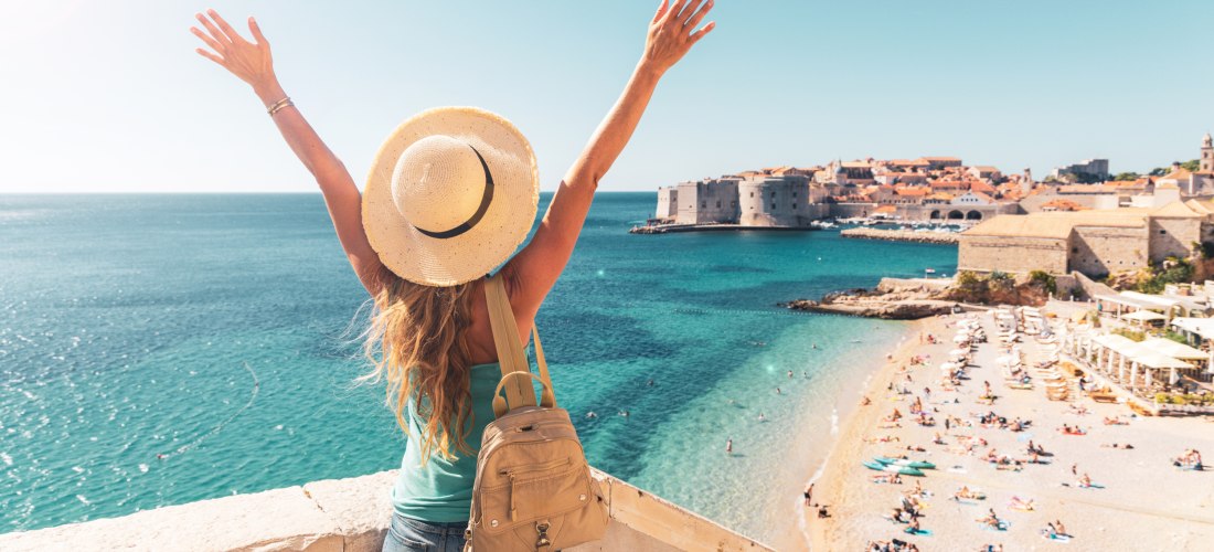 Eine Frau mit Strohhut, Rucksack und sommerlicher Kleidung steht mit erhobenen Armen auf einer Aussichtsterrasse und blickt auf das türkisfarbene Meer und die Altstadt von Dubrovnik. Unter ihr liegt ein belebter Sandstrand mit zahlreichen Sonnenanbetern. 
