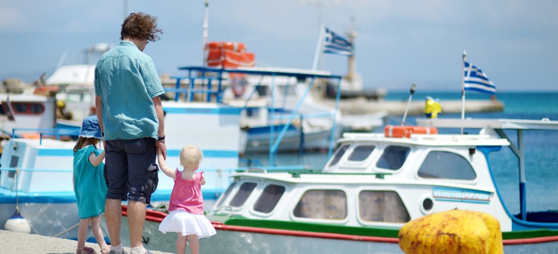 Ein Mann steht mit zwei kleinen Kindern an einem Hafen mit griechischen Fischerbooten. Die Gruppe ist von hinten zu sehen. Die Kinder halten seine Hände – das eine trägt ein blaues Kleid und einen Sonnenhut, das andere ein pinkes Top mit weißem Rock. Im H