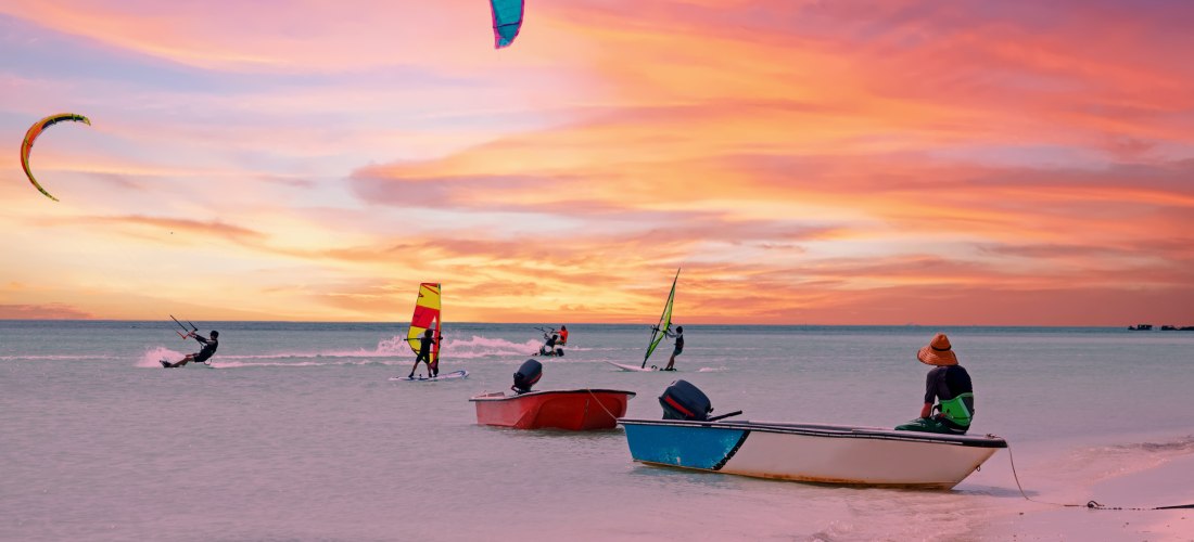 Mehrere Personen beim Kitesurfen und Windsurfen auf ruhigem Wasser vor Aruba bei Sonnenuntergang. Der Himmel leuchtet in warmen Farben von Orange bis Lila. Drei kleine Boote liegen am Ufer, eine Person sitzt auf einem Boot und beobachtet das Geschehen.
