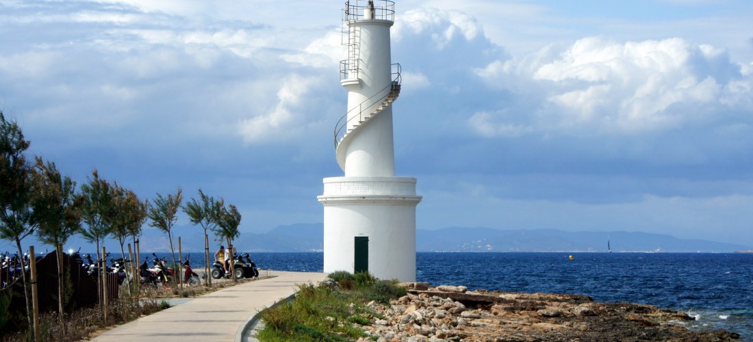 Weißer Leuchtturm mit spiralförmiger Außentreppe an einem Küstenweg, umgeben von Felsen, Büschen und dem Meer. Der Himmel ist leicht bewölkt, im Hintergrund sind Berge zu sehen.