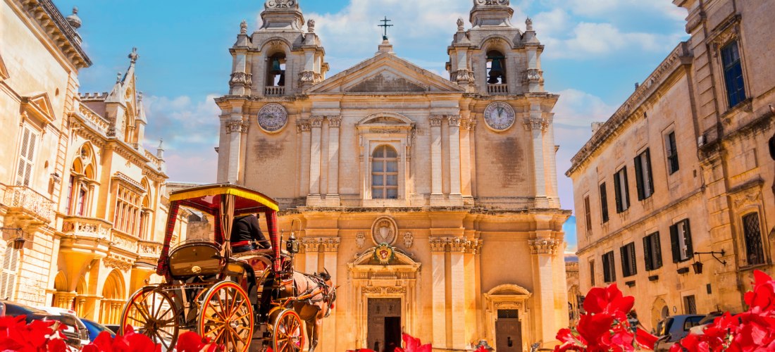 Barocke Kathedrale St. Paul in Mdina auf Malta mit Pferdekutsche im Vordergrund und roten Blumen unter blauem Himmel