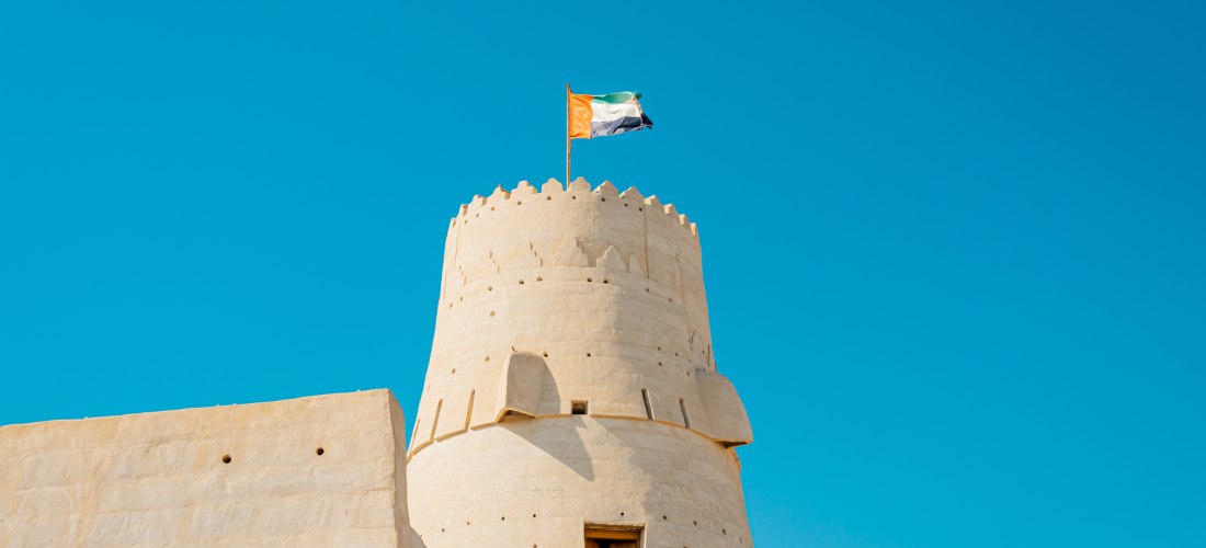 Rundturm des Nationalmuseums Ras Al Khaimah mit Flagge der VAE vor blauem Himmel.