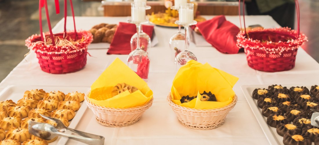 Ein festlich gedeckter Tisch mit verschiedenen traditionellen Keksen und Plätzchen, darunter helles Gebäck, mit Schokolade überzogene Kekse und zwei kleine Körbe mit gelben Servietten. Im Hintergrund rote Dekokörbchen, Gläser mit Kerzen und Geschirr – all