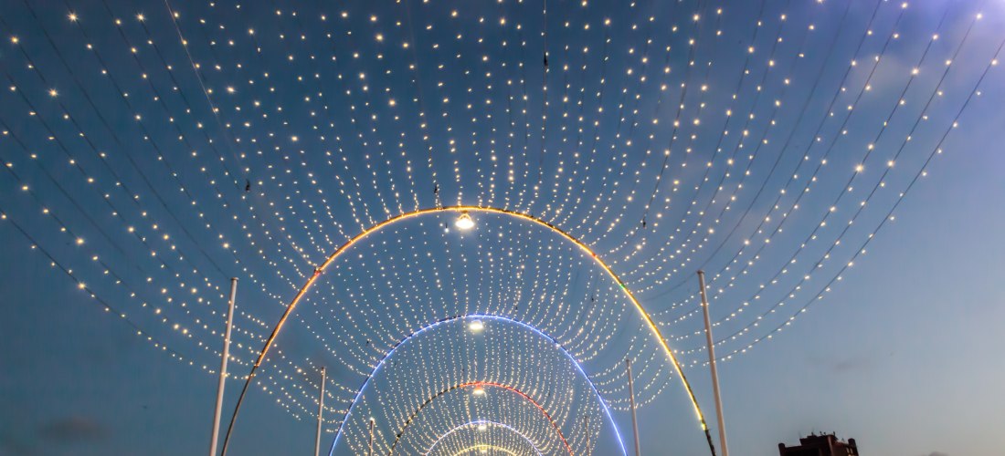 mma-Brücke in Willemstad, Curaçao, am AbeDie berühmte Königin-End. Über der Brücke spannen sich mehrere Bögen aus leuchtenden Lichterketten. Menschen spazieren unter dem funkelnden Lichterhimmel hindurch. Am Rand sind bunte Häuserfassaden zu erkennen