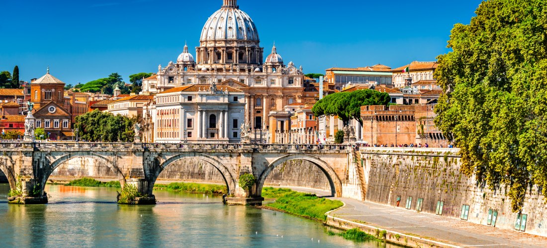Blick über den Fluss Tiber auf den Petersdom im Vatikan mit seiner imposanten Kuppel. Davor die historische Steinbrücke Ponte Sant’Angelo mit Statuen auf den Pfeilern. Die Szene wird eingerahmt von antiken Gebäuden, Zypressen und einem klaren, tiefblauen 