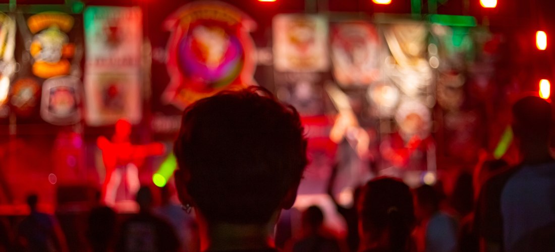 Verschwommene Aufnahme eines Konzert- oder Festivalabends mit bunten Lichtern. Im Vordergrund steht eine Person mit dem Rücken zur Kamera, die auf die hell erleuchtete Bühne mit Live-Musik blickt.