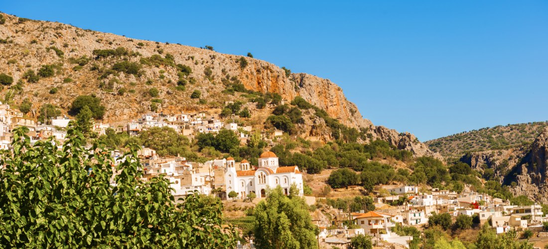 Weißes mediterranes Dorf mit Kirche an einem felsigen Berghang. Die Kirche mit roten Kuppeln ist von traditionellen Häusern und spärlicher Vegetation umgeben, unter einem klaren blauen Himmel