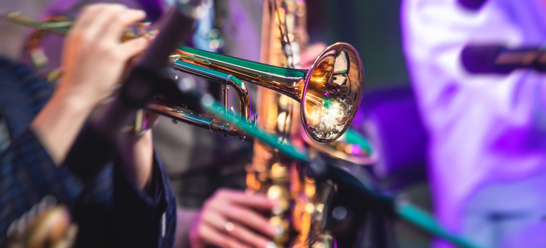 Nahaufnahme von Musikern, die auf Trompeten und Saxophonen spielen. Die Szene ist farbenfroh beleuchtet, Instrumente glänzen im Licht.