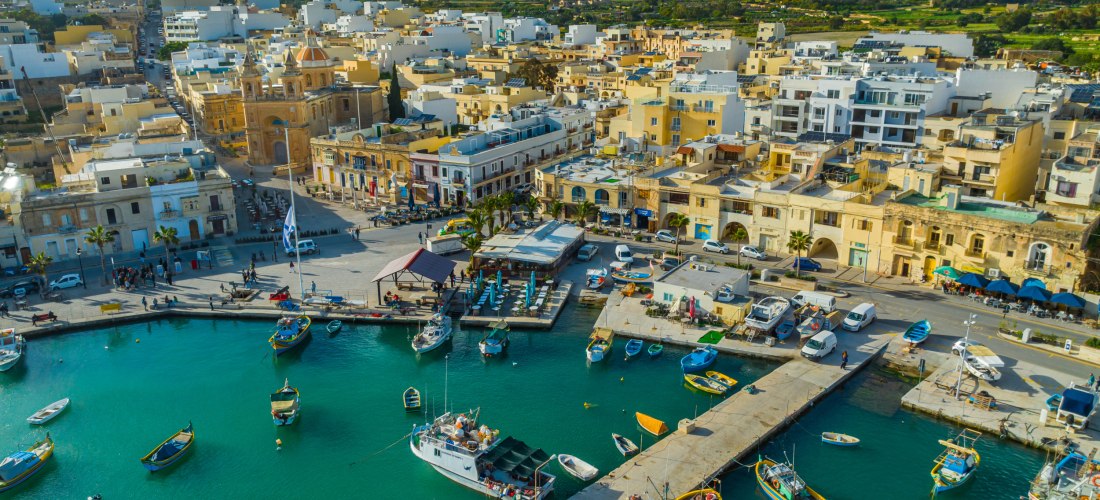 Luftaufnahme des Hafens von Marsaxlokk auf Malta mit bunten Fischerbooten, Promenade und historischen Gebäuden im Hintergrund