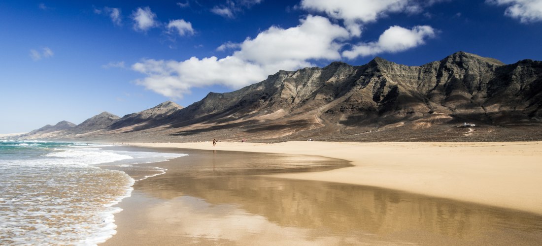 Weitläufiger, menschenleerer Sandstrand mit sanften Wellen. Im Hintergrund eine zerklüftete Gebirgskette unter blauem Himmel mit vereinzelten Wolken