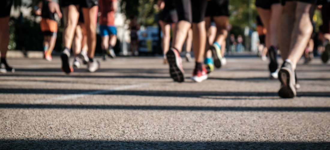 Nahaufnahme von Beinen und Laufschuhen mehrerer Teilnehmer eines Straßenlaufs. Die Szene zeigt Bewegung und Dynamik auf asphaltierter Straße bei sonnigem Wetter