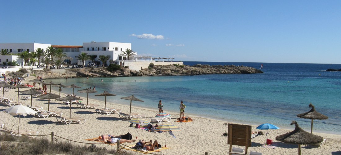 Sandstrand von Es Pujols auf Formentera mit Liegen, Sonnenschirmen aus Stroh, Urlaubern am Strand und türkisblauem Meer. Im Hintergrund weiße Gebäude mit Palmen direkt an der Küste.
