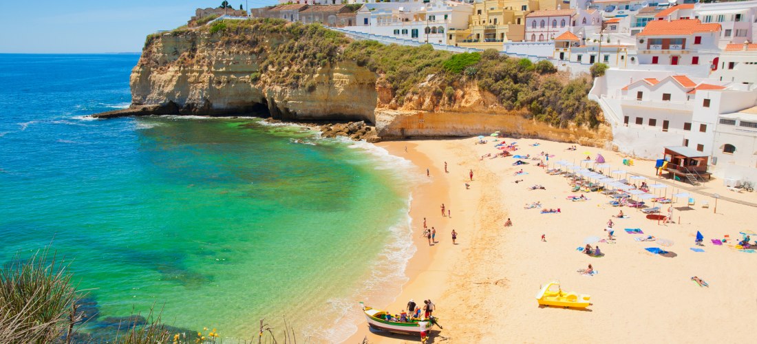 Algarve-Bucht mit goldgelbem Sandstrand und türkisgrünem Wasser. Über den hellen Klippen stehen weiß getünchte Häuser; am Strand baden und entspannen Menschen.