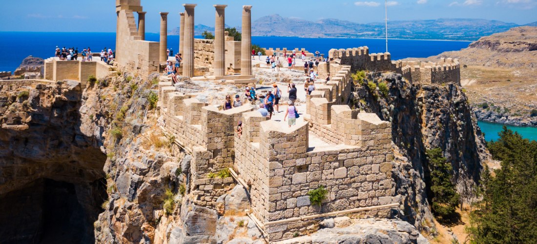 Das Bild zeigt die Akropolis von Lindos auf der griechischen Insel Rhodos, eine gut erhaltene antike Burganlage auf einem steilen Felsplateau.   Die Festung besteht aus massiven, hellen Steinmauern mit Zinnen, Treppen und historischen Säulen, von denen ei