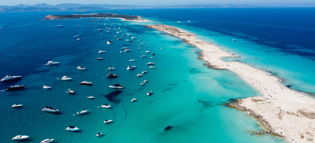 Luftaufnahme einer langen, schmalen Sandbank in Formentera, Spanien. Zahlreiche weiße Boote und Yachten ankern im türkisblauen, klaren Wasser auf beiden Seiten der Landzunge. Die Szenerie erstreckt sich bis zum Horizont, wo sanfte Hügel und das offene Mee