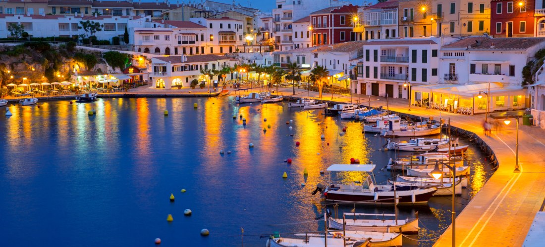 Hafen von Mahón auf Menorca in der Abenddämmerung. Boote liegen am Kai, die Uferpromenade und Häuser sind stimmungsvoll beleuchtet.