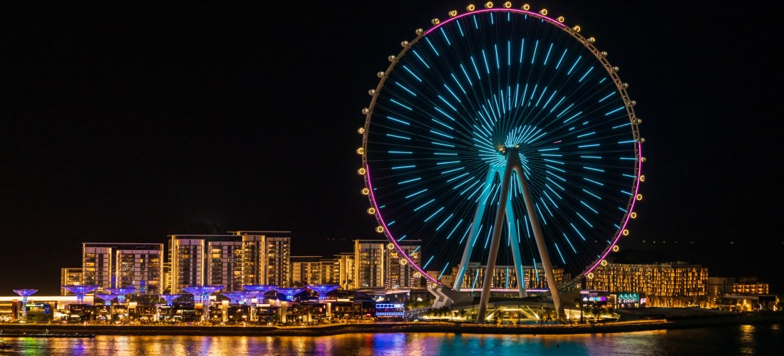 Dubai - Bluewaters Island Das beleuchtete Riesenrad Ain Dubai auf Bluewaters Island bei Nacht. Die Lichtinstallation am Rad leuchtet in Blau- und Lilatönen. Im Vordergrund spiegelt sich das Licht auf dem Wasser, dahinter sind moderne Gebäude und Restaurants stimmungsvoll beleuchte