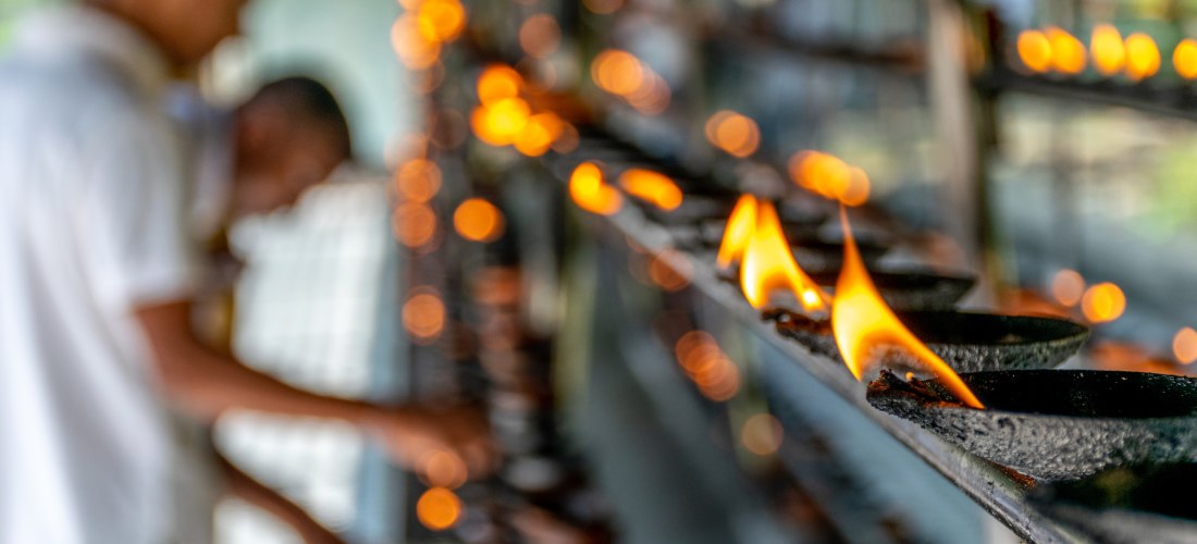 Reihe kleiner Ton-Öllampen mit offenen Flammen auf einer Metallablage im Tempel. Im Hintergrund sind unscharf zwei Menschen zu sehen, die vermutlich weitere Lampen entzünden. Das Bild vermittelt eine ruhige, spirituelle Atmosphäre.
