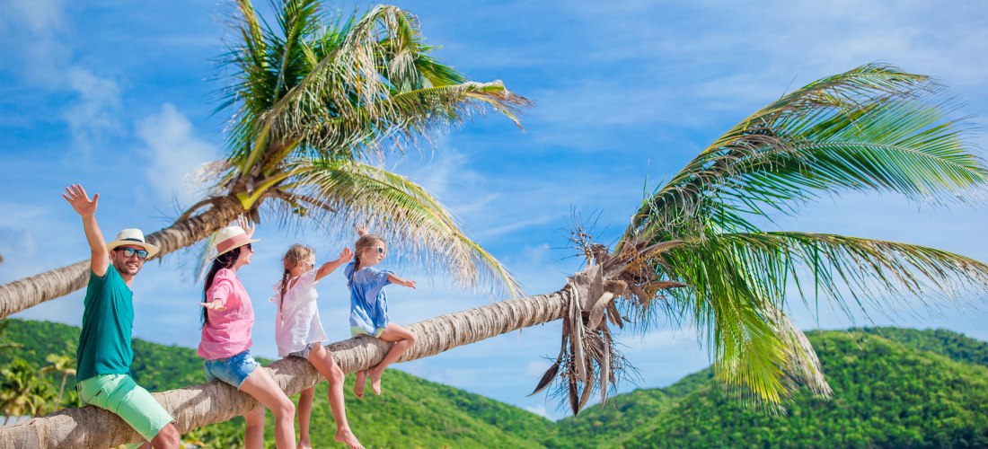 Fröhliche Familie sitzt auf einer geneigten Palme an einem tropischen Strand mit kristallklarem Wasser und grünen Hügeln im Hintergrund