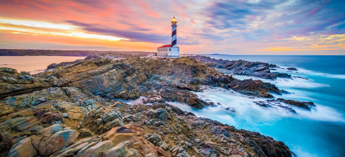 Stimmungsvolle Küstenlandschaft bei Sonnenuntergang auf Menorca, Spanien. Im Mittelpunkt steht der rot-weiß gestreifte Leuchtturm Faro de Favàritx auf einer schroffen Felsklippe. Die Felsen sind bizarr geformt und ziehen sich entlang der wilden Küste. Der