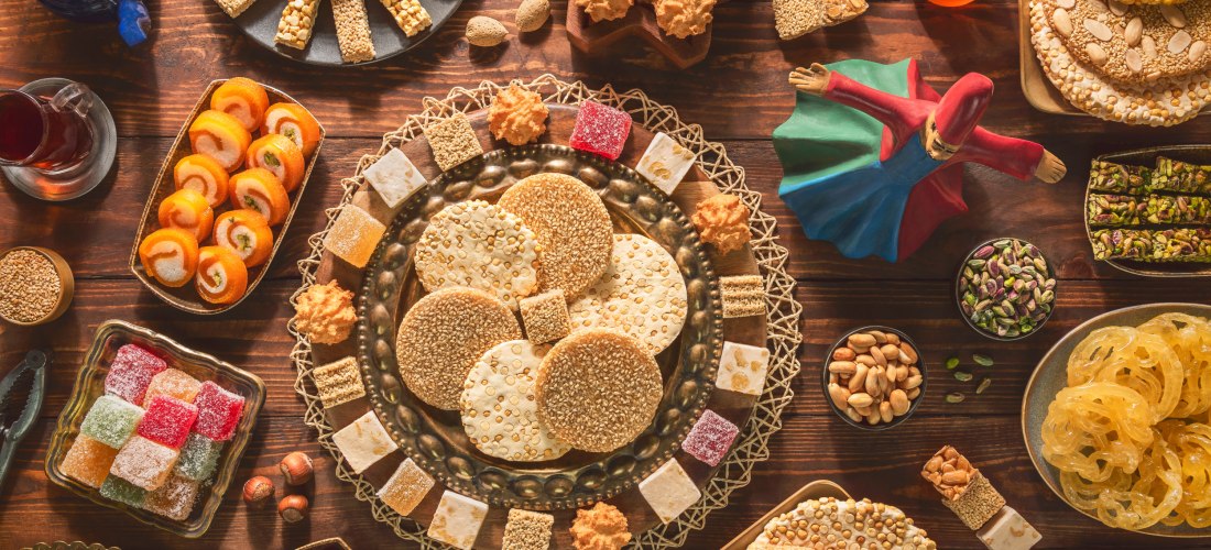 Vielfältige orientalische Süßigkeiten auf einem dunklen Holztisch, darunter Sesamkekse, Türkischer Honig, Nüsse, Gebäck und Lokum in verschiedenen Farben. In der Mitte ein kunstvoll gedeckter Teller, daneben eine Figur eines tanzenden Derwischs