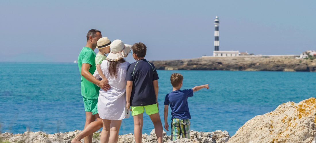 Eine Familie mit fünf Personen steht barfuß auf einem Felsen an der Küste und blickt auf das Meer. Die Erwachsenen tragen Sommerkleidung und Hüte, ein Kind zeigt auf einen Leuchtturm in der Ferne. Der Leuchtturm ist schwarz-weiß gestreift und steht auf ei