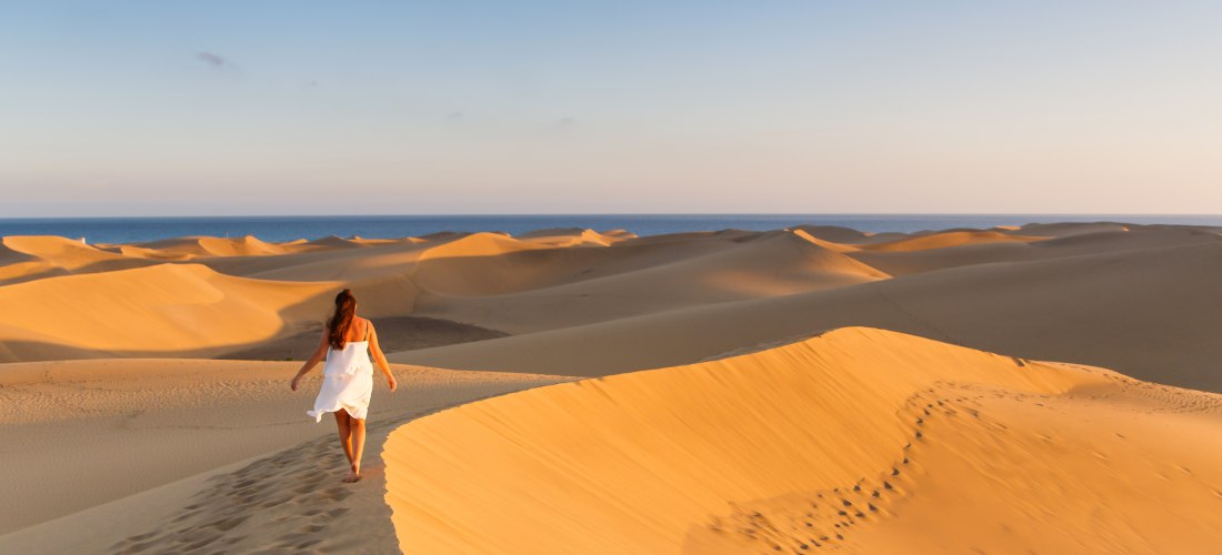 Frau in weißem Kleid geht durch die goldgelben Dünen von Maspalomas auf Gran Canaria, im Hintergrund das Meer unter blauem Himmel.