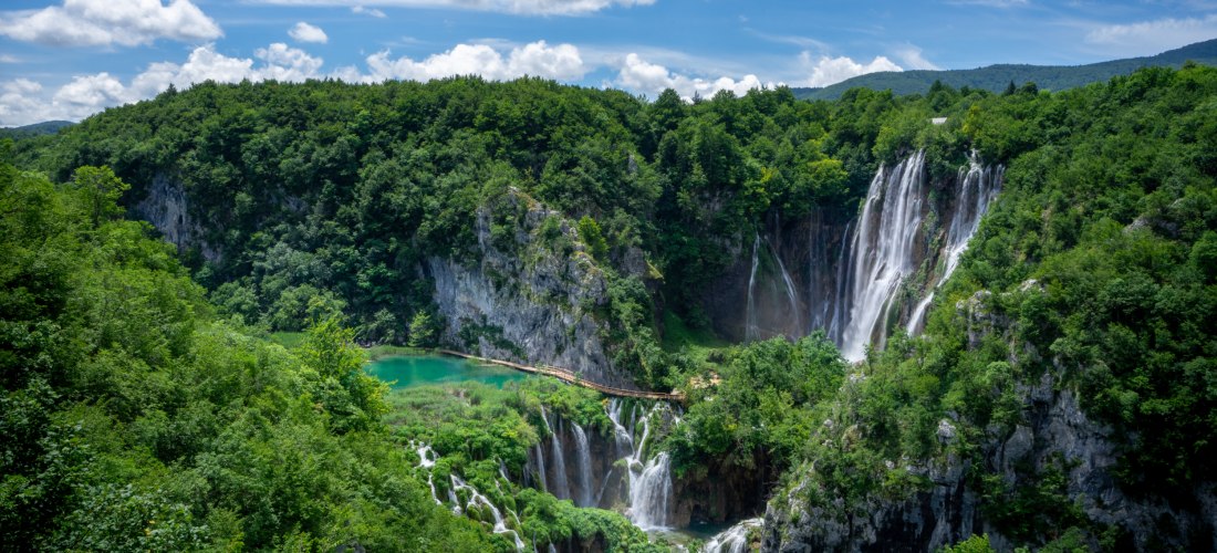 Panoramablick auf die Plitvicer Seen in Kroatien. Mehrere Wasserfälle stürzen von bewaldeten Felswänden in türkisfarbene Seen, die durch Holzstege miteinander verbunden sind. Der Himmel ist blau mit einigen weißen Wolken.