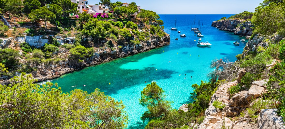Eine idyllische Bucht auf Mallorca mit klarem, türkisblauem Wasser, gesäumt von grünen Pinien und steilen Klippen. Segelboote und Yachten schwimmen im Wasser, während sich einige Menschen in der Bucht entspannen. Rechts im Bild windet sich ein schmaler Pf