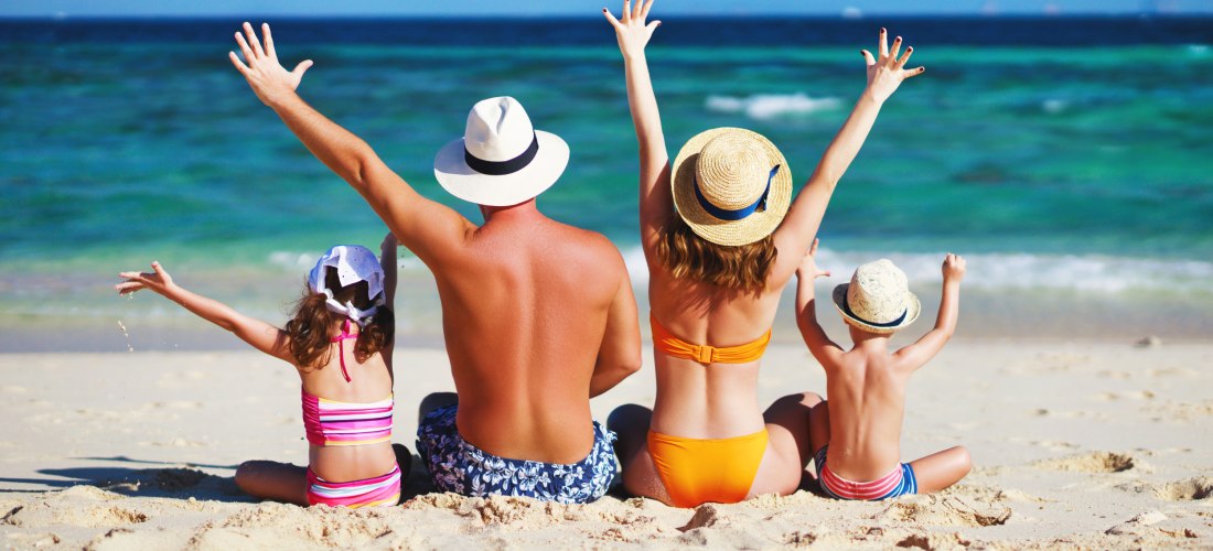 Eine Familie mit zwei Kindern sitzt am Strand und schaut aufs Meer. Alle tragen Badebekleidung und Sonnenhüte, die Arme sind fröhlich in die Luft gestreckt. Die Stimmung ist entspannt und sommerlich.