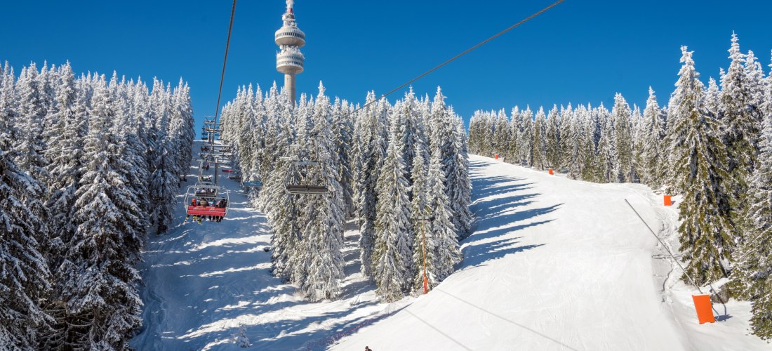 Winterlandschaft im Skigebiet Pamporovo in Bulgarien. Ein Sessellift fährt durch verschneite Tannenwälder hinauf zu den Pisten, während ein einzelner Skifahrer die weiße Piste hinuntergleitet. Im Hintergrund erhebt sich der markante Fernsehturm Snezhanka 