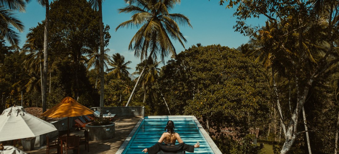 Eine Frau in schwarzer Badebekleidung steht am Rand eines Infinity-Pools, der von Palmen und dichtem tropischem Grün umgeben ist. Sie schaut in die Ferne, die Arme leicht ausgebreitet. Im Hintergrund stehen Sonnenschirme und Holzmöbel auf der Terrasse. Di