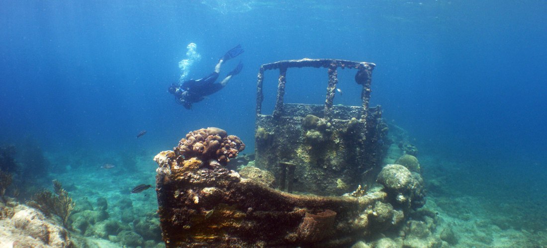 Taucher erkundet ein versunkenes Schiffswrack unter Wasser. Das Wrack ist teilweise mit Korallen bewachsen und liegt auf einem felsigen Meeresboden. Im klaren blauen Wasser sind einige Fische zu sehen, während Sonnenlicht durch die Wasseroberfläche dringt