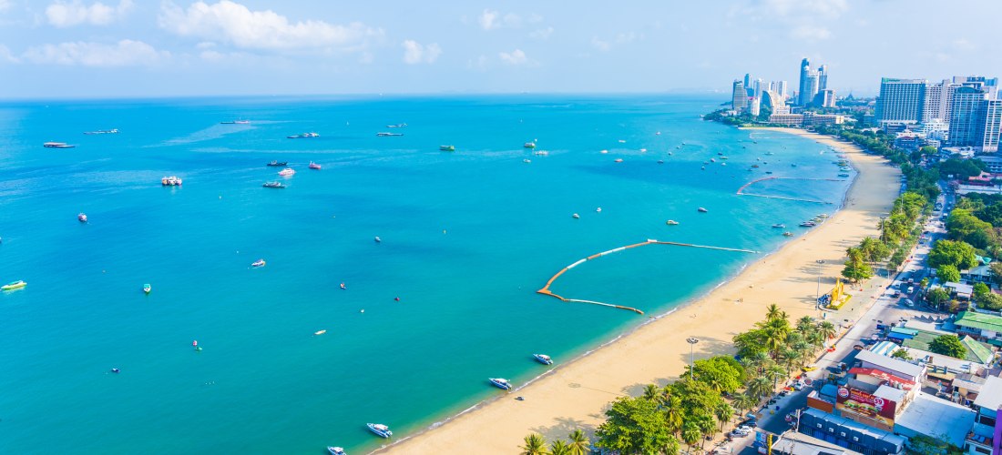 Luftaufnahme von Pattaya Beach mit goldenem Sandstrand, türkisblauem Meer und zahlreichen Booten. Entlang der Küste reihen sich Hochhäuser, Hotels und eine lebhafte Uferstraße.